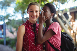 Female wearing a backpack whispers to another female who is smiling at the camera.