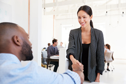 Standing female shakes the hand of a seated male in a surgical white office space.