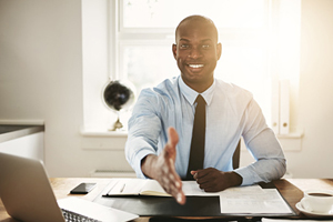 Male of color extending his hand for the camera to shake as he sits at an office desk.