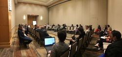 Photo of an event space with beige walls and chairs arranged around the edges of the room with a few dozen people sitting.