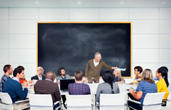 Teach pointing at a student while standing in front of a black board at the front of a classroom.
