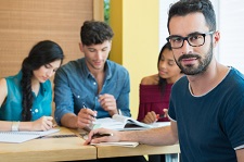 Male in glasses looking at the camera as three others study together in the background.