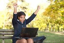 Female of color raising both her arms in the air in an act of celebration while using her laptop outside on a park bench.