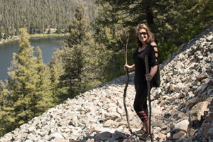 female holds two sticks, one in either hand, while posing on a hill overlooking a body of water and trees