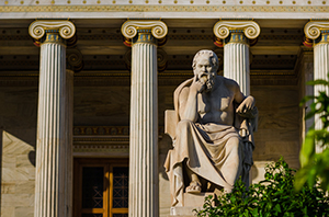grey building with large marble columns visible in the background and a statue of a bearded man in a cloth stroking his chin as though deep in thought