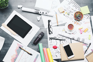 cluttered desk with a range of items such as glasses, pens, papers, smartphone, and more scattered