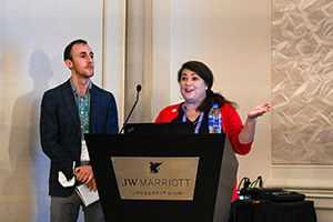 female in a red blouse speaks from behind a podium while a male in a green collared shirt and blazer stands next to her