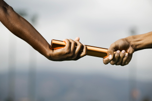 Hands passing a small wooden block between people outdoors, symbolizing trust or cooperation