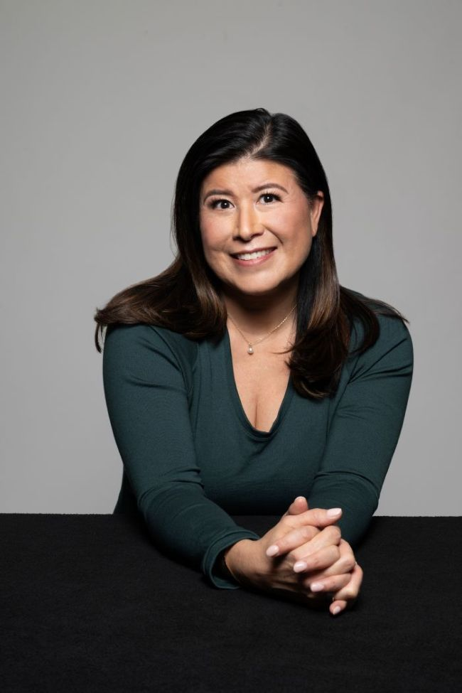 Portrait of a smiling woman seated at a table against a plain gray background