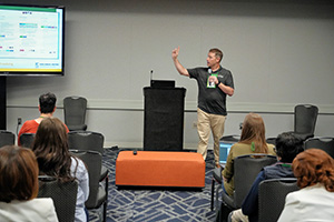 A presenter teaching a class in a conference room with an audience seated and a screen showing slides