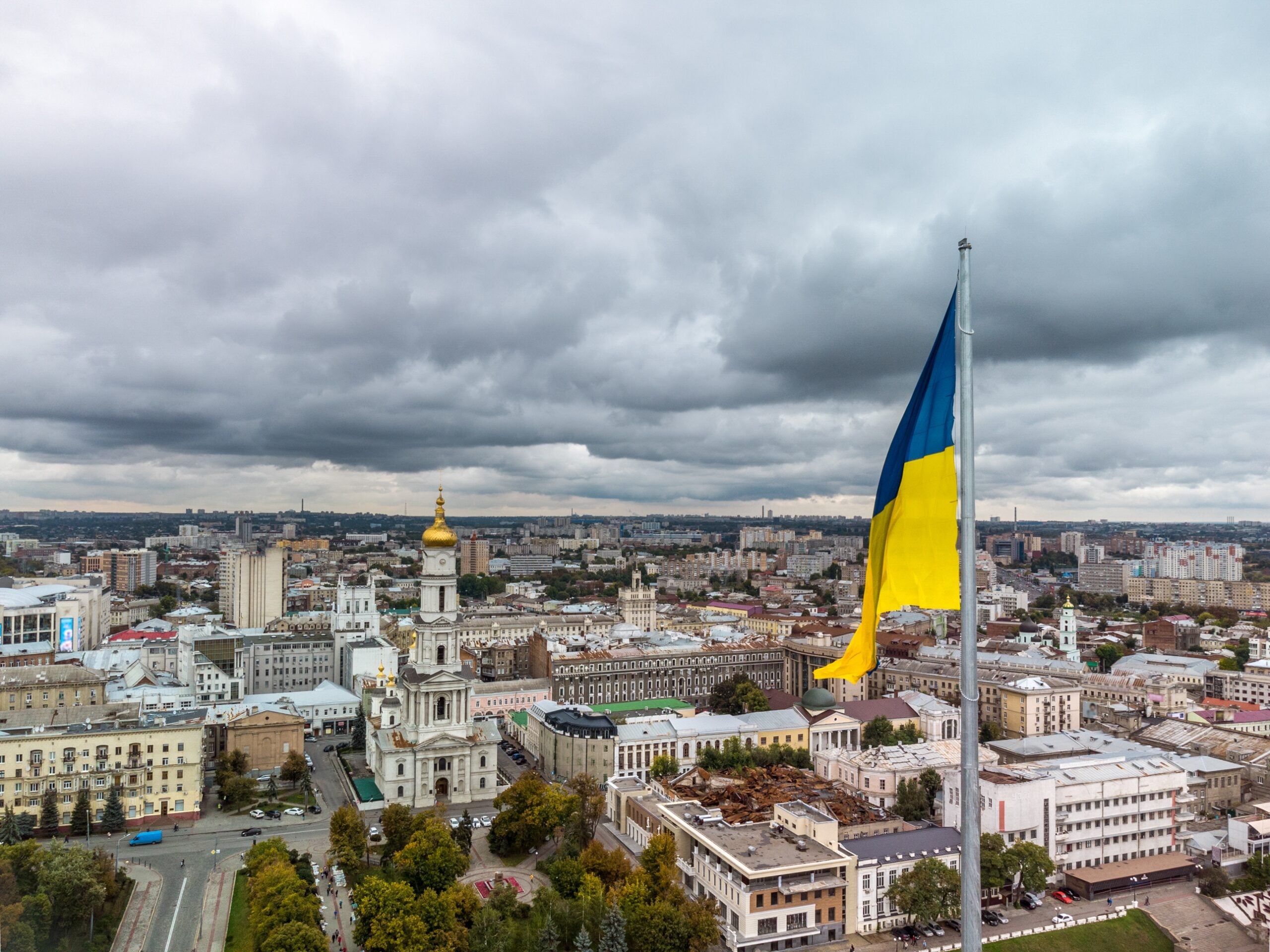
The Ukrainian flag flying over a city skyline under a cloudy sky.