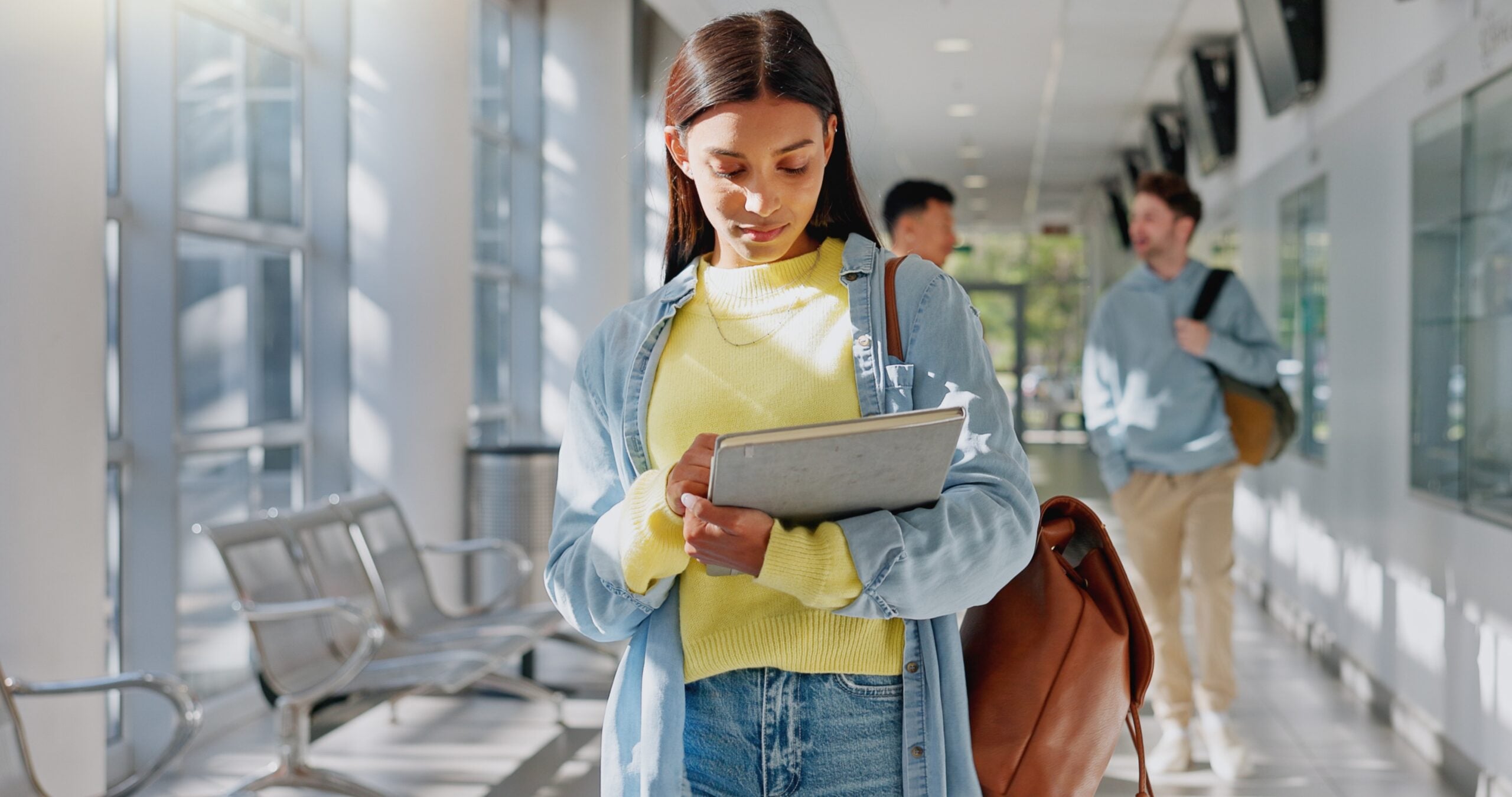 
A student walking through a university hallway while looking down at a tablet.