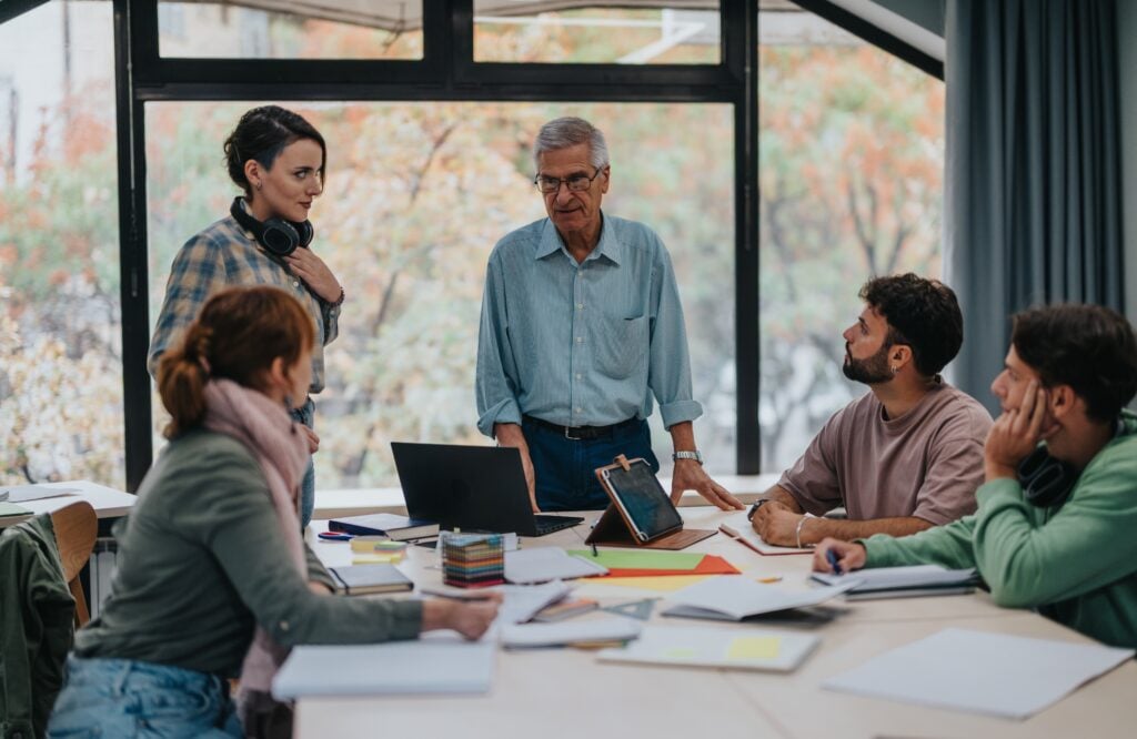 Claude responded: An older man leading a discussion with a small group around a table in a bright meeting room.An older man leading a discussion with a small group around a table in a bright meeting room