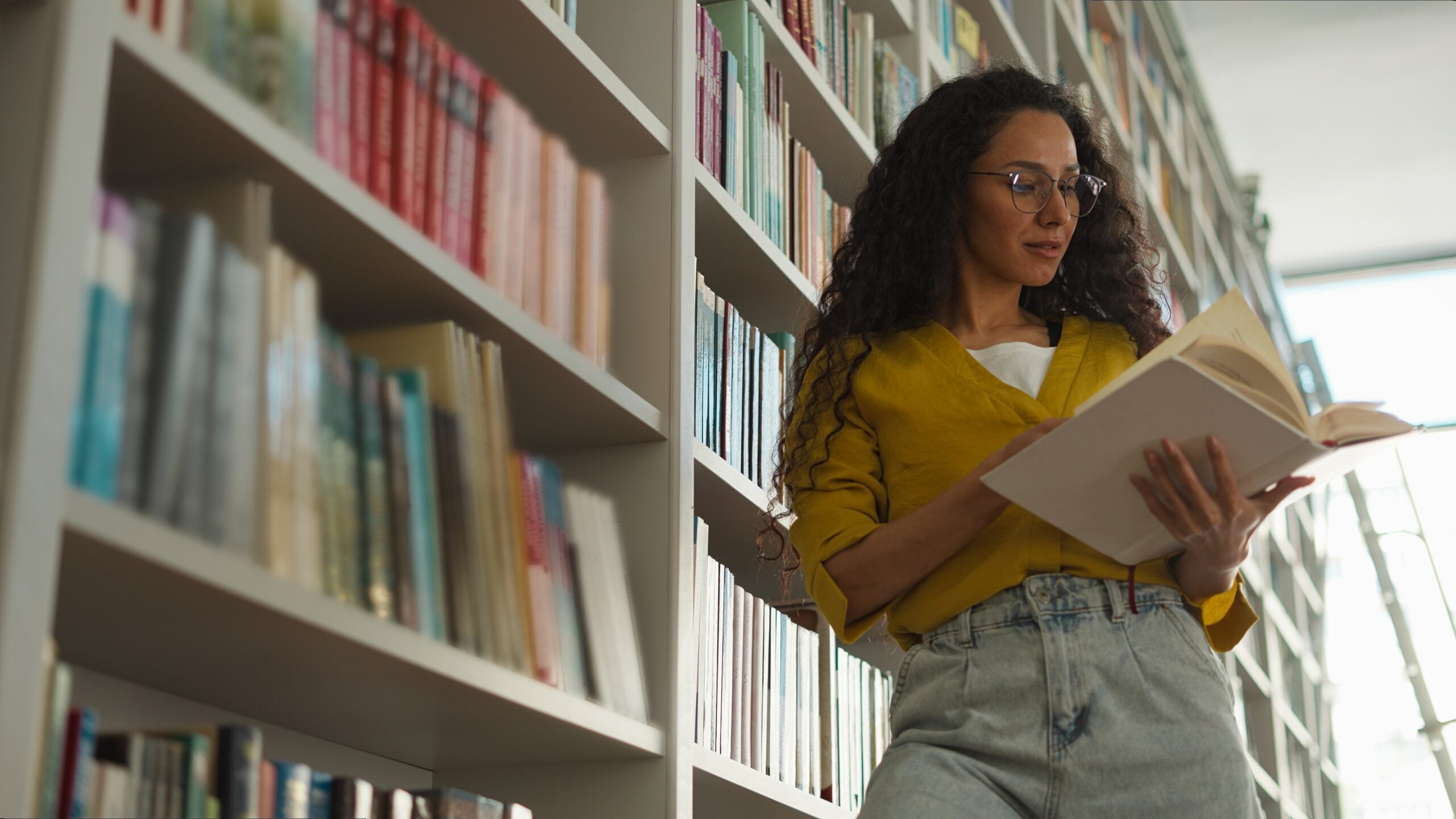 A woman reading a book beside shelves in a library.