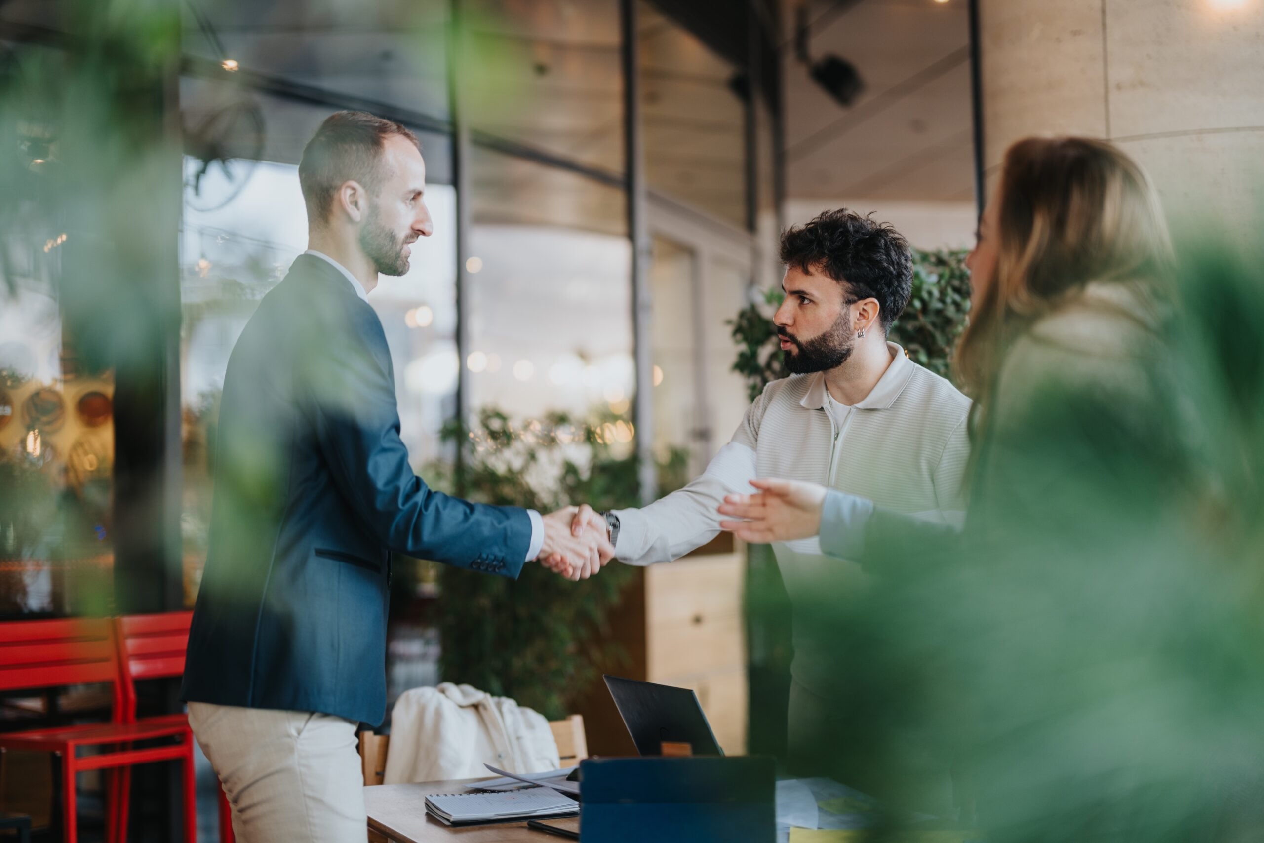 Two men shaking hands in a modern workspace, with a woman nearby.
