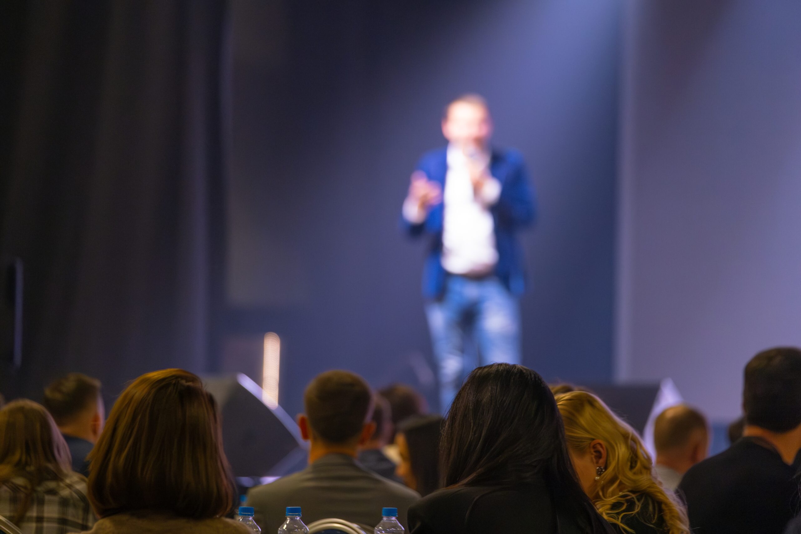 A speaker on stage addresses an audience at a conference or event.