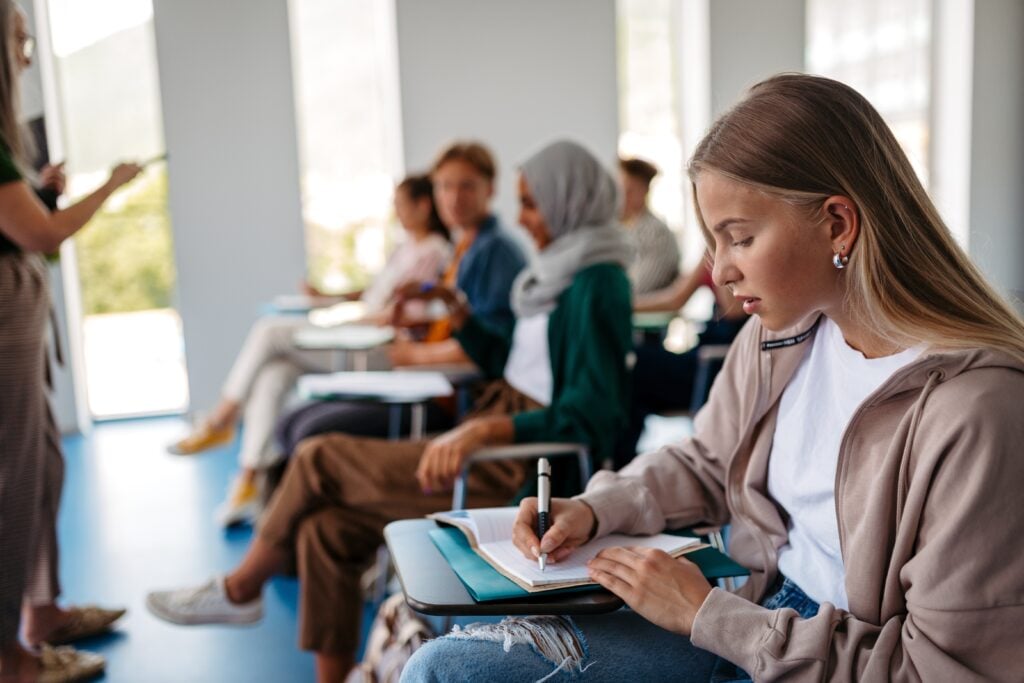 Claude responded: A student taking notes at a desk in a bright classroom.A student taking notes at a desk in a bright classroom.