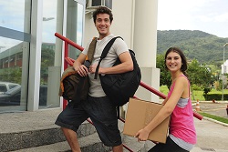 Two people smiling as they carry bags and a box up some stairs.
