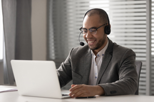 male smiles on a video call on their laptop while wearing a mic and headphones