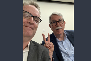 two businessmen pose for a selfie together as one holds up the two-finger peace sign