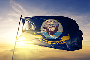 Photo of the United States Navy flag flying in the wind with a sunny sky in the background.