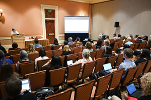 audience looking at a projector screen as they sit in brown backed chairs