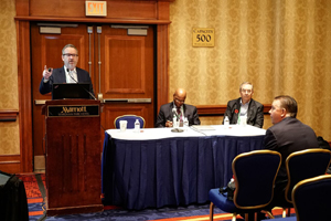 male speaks from behind a podium while 2 other males sit at a panelist table to the speaker's left