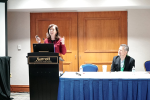 female presents from a podium with a male sitting at a panelist table to her left