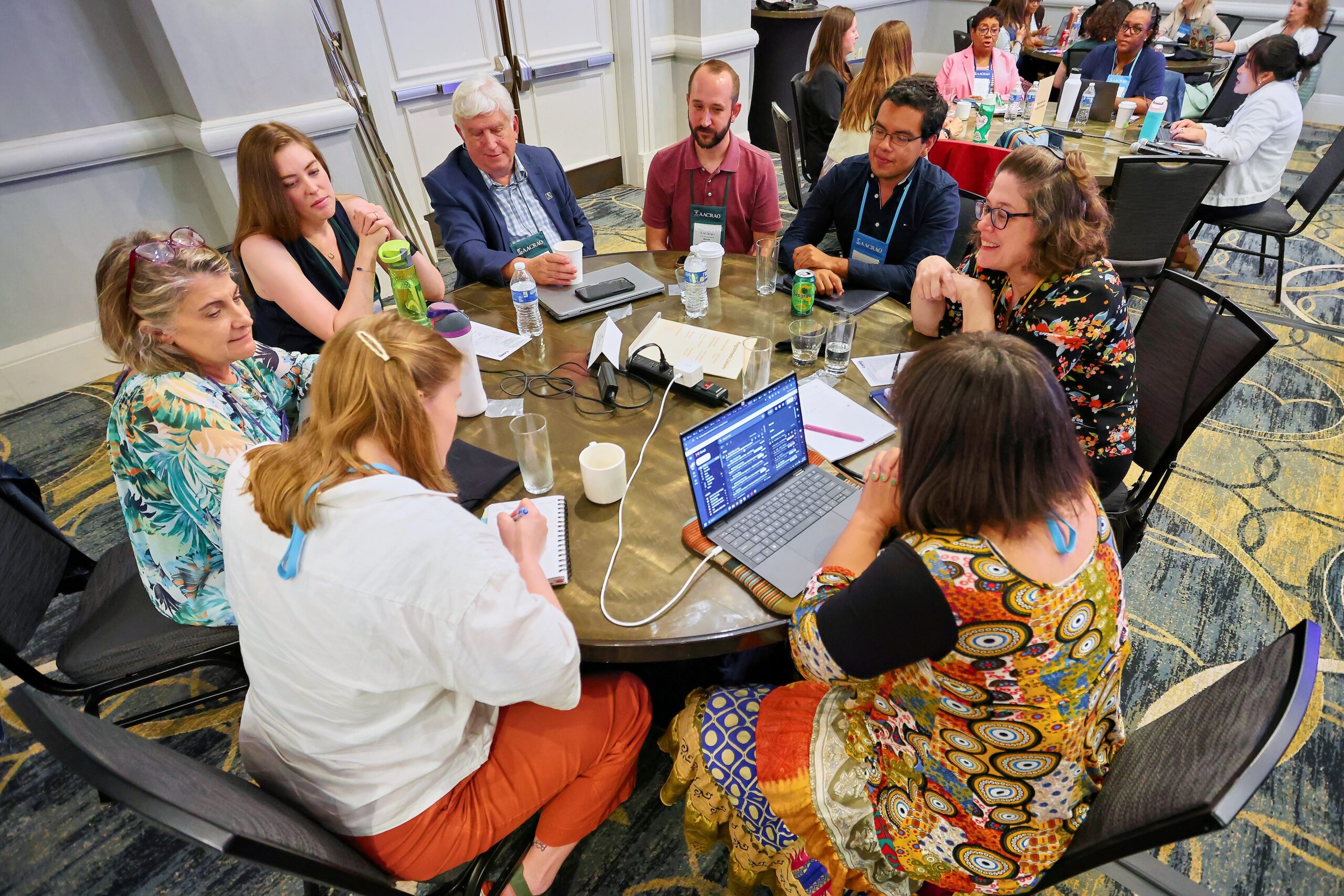 Group of six people working at a round table talking and taking notes on laptops